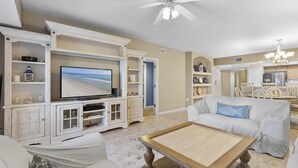 Ein Wohnzimmer mit Fernseher, weißen Möbeln, einem Deckenventilator und einer Küche im Hintergrund.