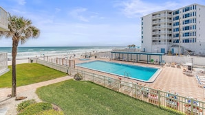 A swimming pool with a view of the ocean, a fenced area, and a building with multiple balconies. - Ocean Reef Villas 207 (New Smyrna Beach)
