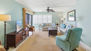 A living room with a sofa, armchair, TV, and a ceiling fan. - Ocean Reef Villas 207 (New Smyrna Beach)