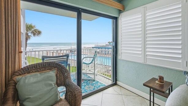 A balcony with a view of the beach, a wicker chair, a small table with a candle, and a white door. - Ocean Reef Villas 207 (New Smyrna Beach)