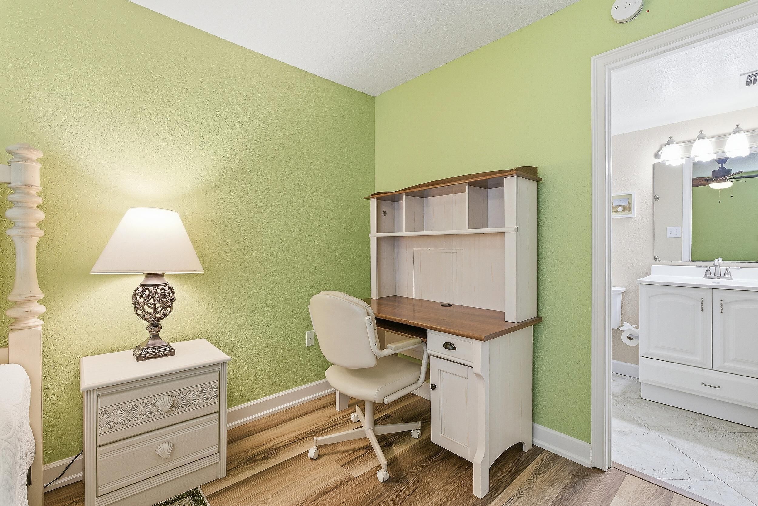 A bedroom with a desk, chair, bedside table, and a bathroom with a mirror and sink.