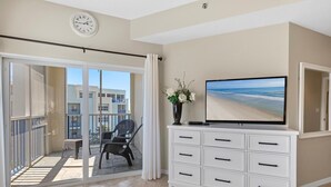 A balcony with a chair and table, a white dresser with drawers, a flat-screen TV, and a clock on the wall. - Oceanwalk 8-507 (New Smyrna Beach)