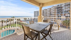 Een balkon met een houten tafel en stoelen, met uitzicht op een zwembad en een strand.