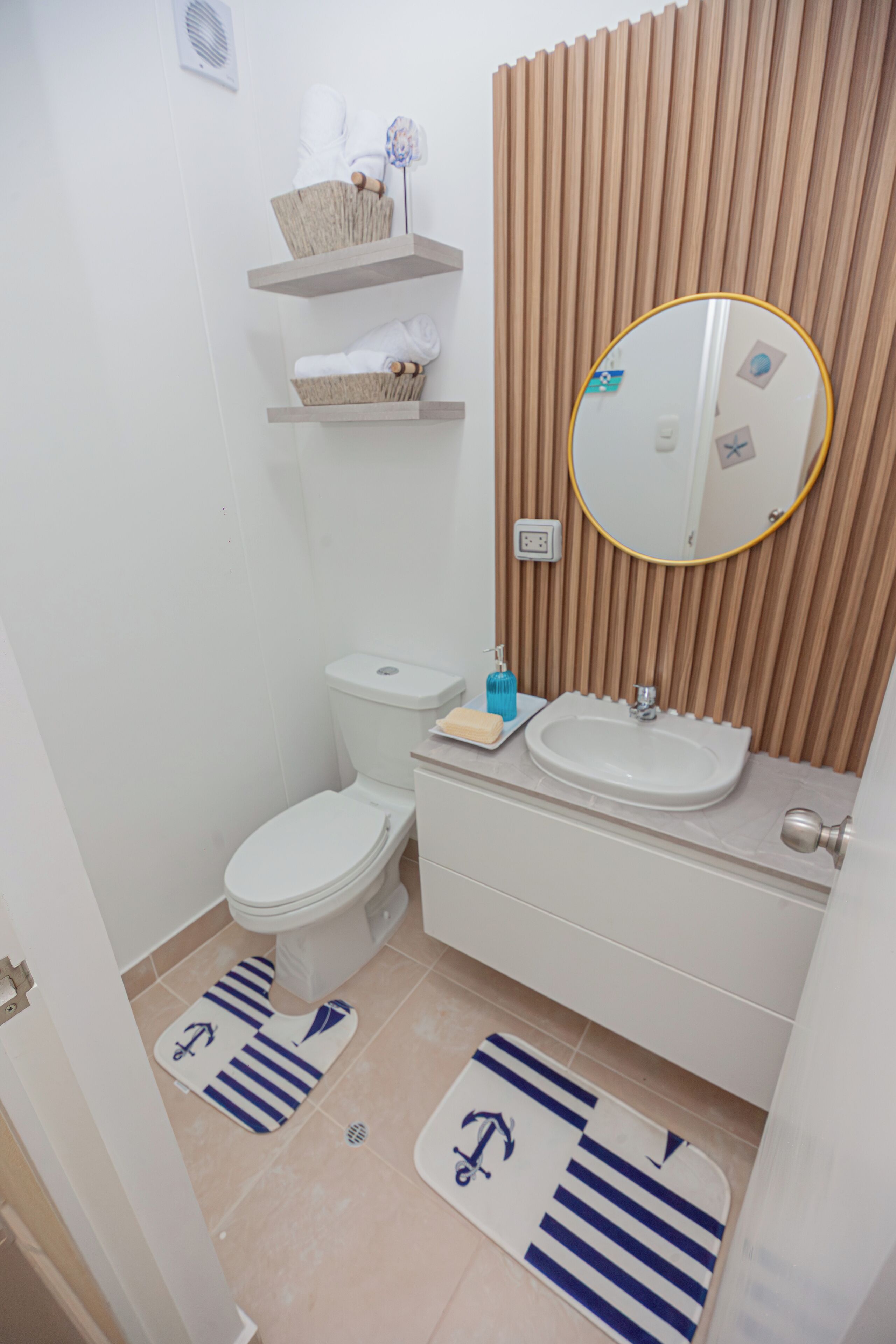 A compact bathroom with a toilet, sink, and mirror. There are two shelves above the sink, one holding a basket and the other a towel. The floor has two striped mats.