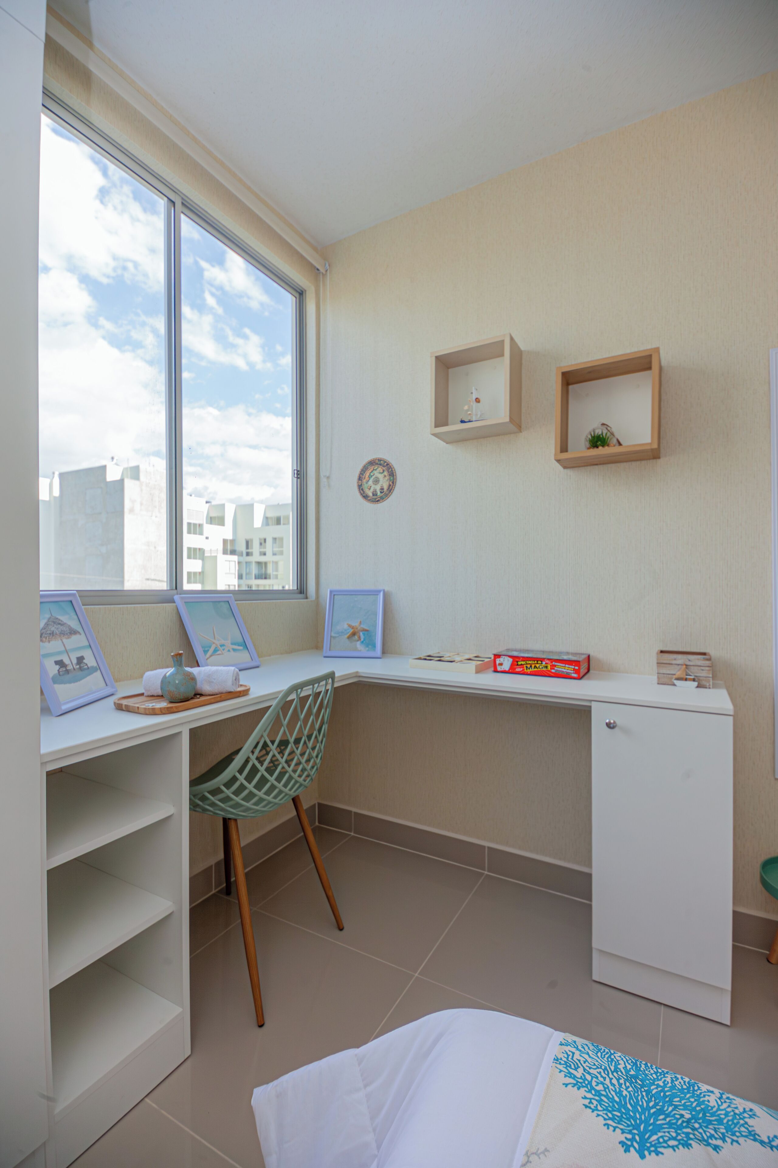Een kleine, goed verlichte kamer met een bureau, stoel en een wandplank. Er is een raam dat natuurlijk licht en uitzicht op gebouwen biedt.