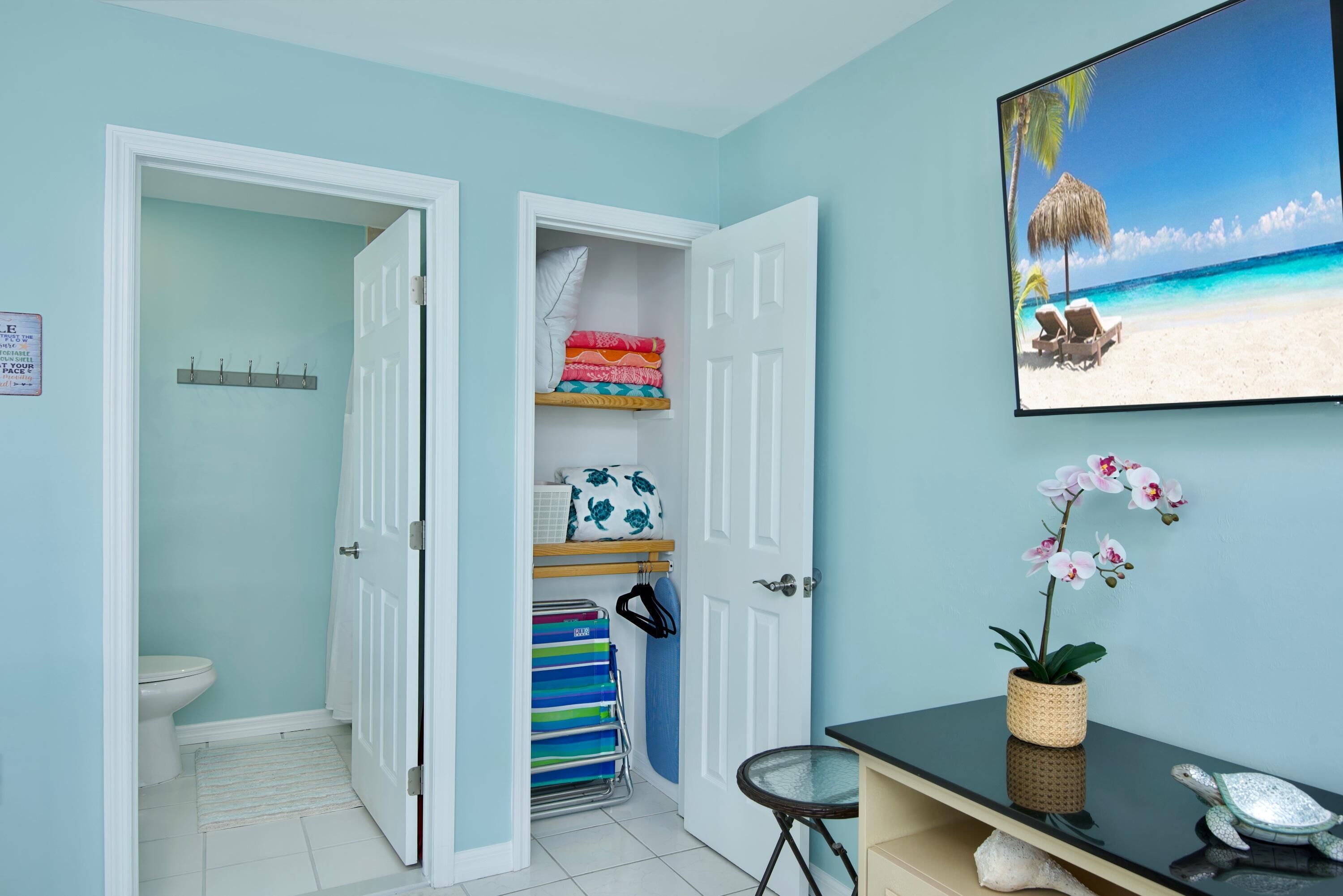 Une salle de bain comprenant des toilettes, une étagère avec des serviettes pliées, un porte-serviettes avec des serviettes colorées et une petite table sur laquelle est posé un objet décoratif.