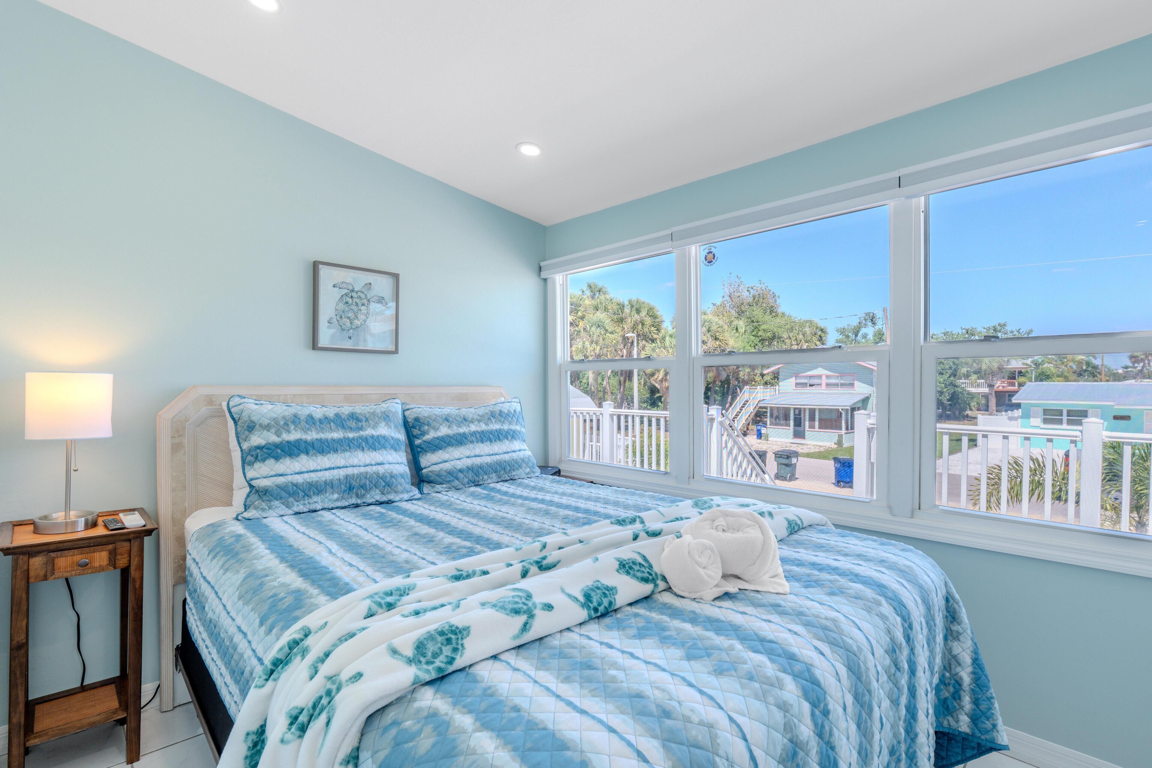 A bedroom with a large window, a bed with a blue and white patterned bedspread, a bedside lamp, and a framed picture on the wall.