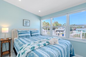 A bedroom with a large window, a bed with a blue and white patterned bedspread, a bedside lamp, and a framed picture on the wall. - Island Paradise 4 (Fort Myers Beach)