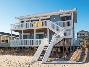 A multi-story beachfront house with a wooden deck and stairs leading to the balconies.