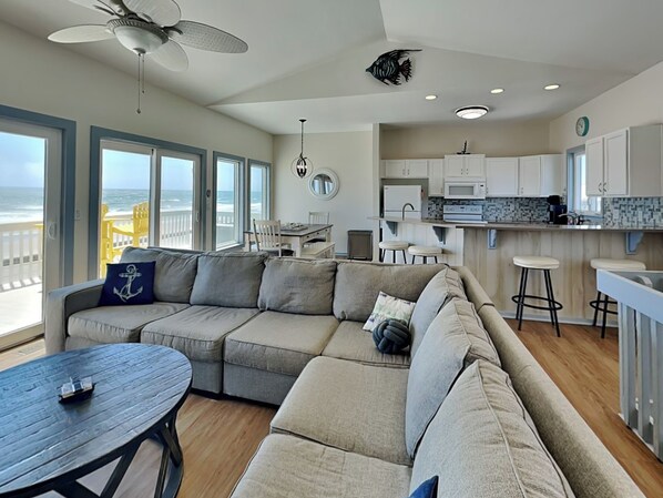 A modern living room with a sectional sofa, a dining table, and a kitchen area. - Sundance by Topsail Realty (Holly Ridge)