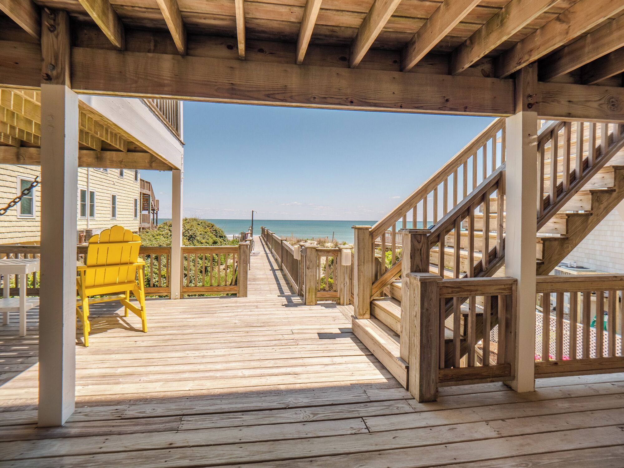 A wooden deck leading to a beach with a yellow chair and a staircase.
