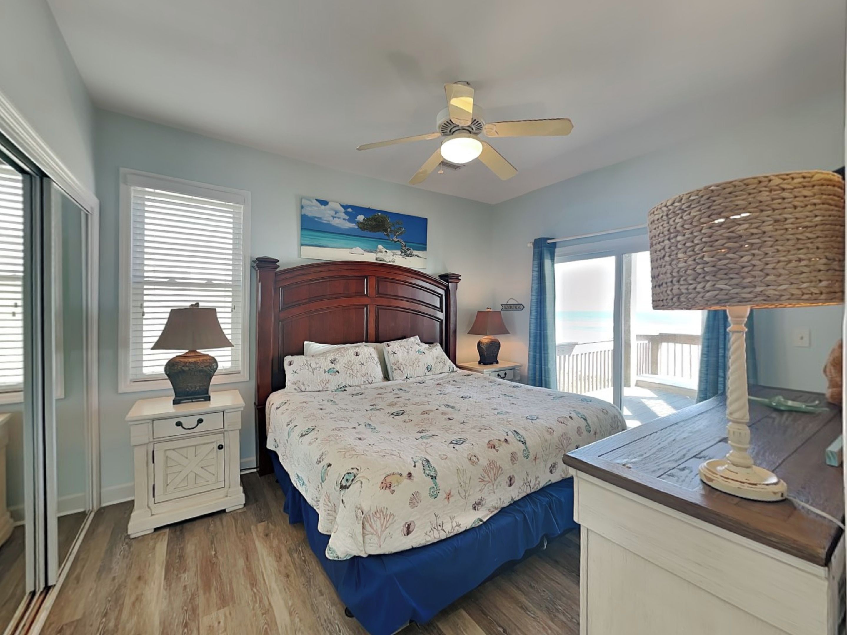 A bedroom with a wooden headboard, a bed with a floral bedspread, a ceiling fan, a nightstand with a lamp, and a view of the ocean through a window.