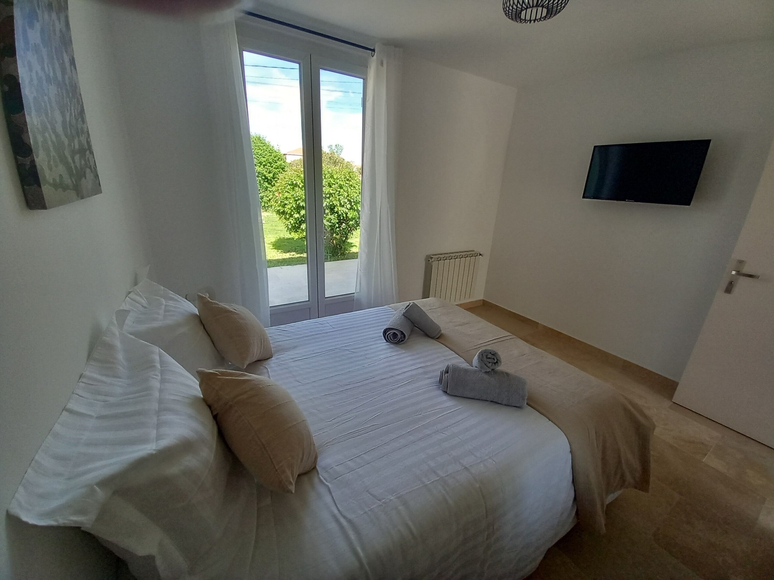 A bedroom with a bed, two pillows, a TV on the wall, and a window with a view of greenery.