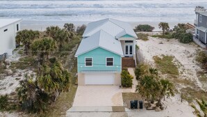 A coastal house with a light blue exterior, a white garage, and a front porch. It is surrounded by sandy beach and palm trees. - Ocean Dream (New Smyrna Beach)