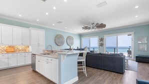 A spacious living area with a large gray sectional sofa, a kitchen with white cabinets and a marble countertop, and a view of the ocean through large windows. - Ocean Dream (New Smyrna Beach)