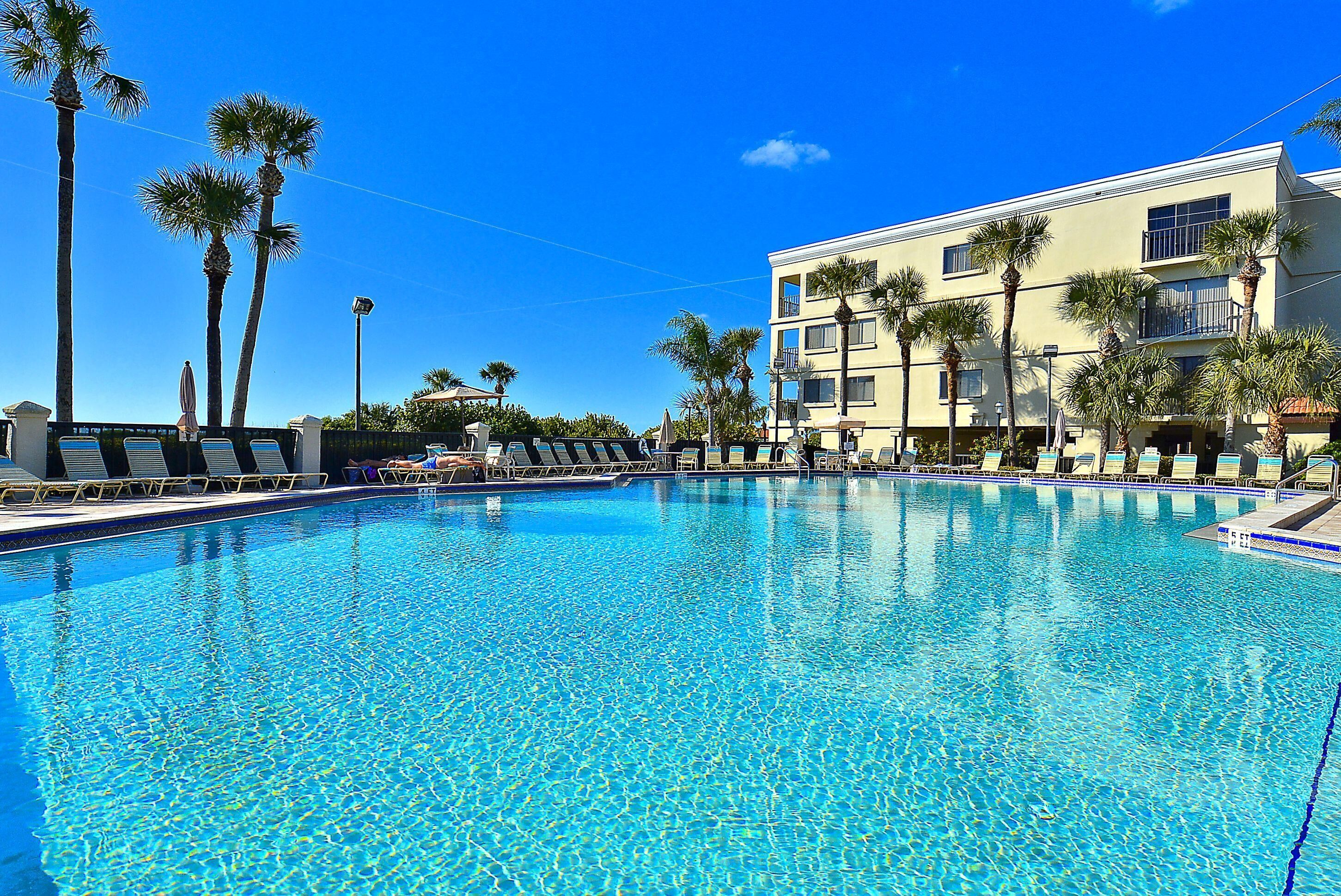 A clear blue pool with lounge chairs, palm trees, and a building with balconies.