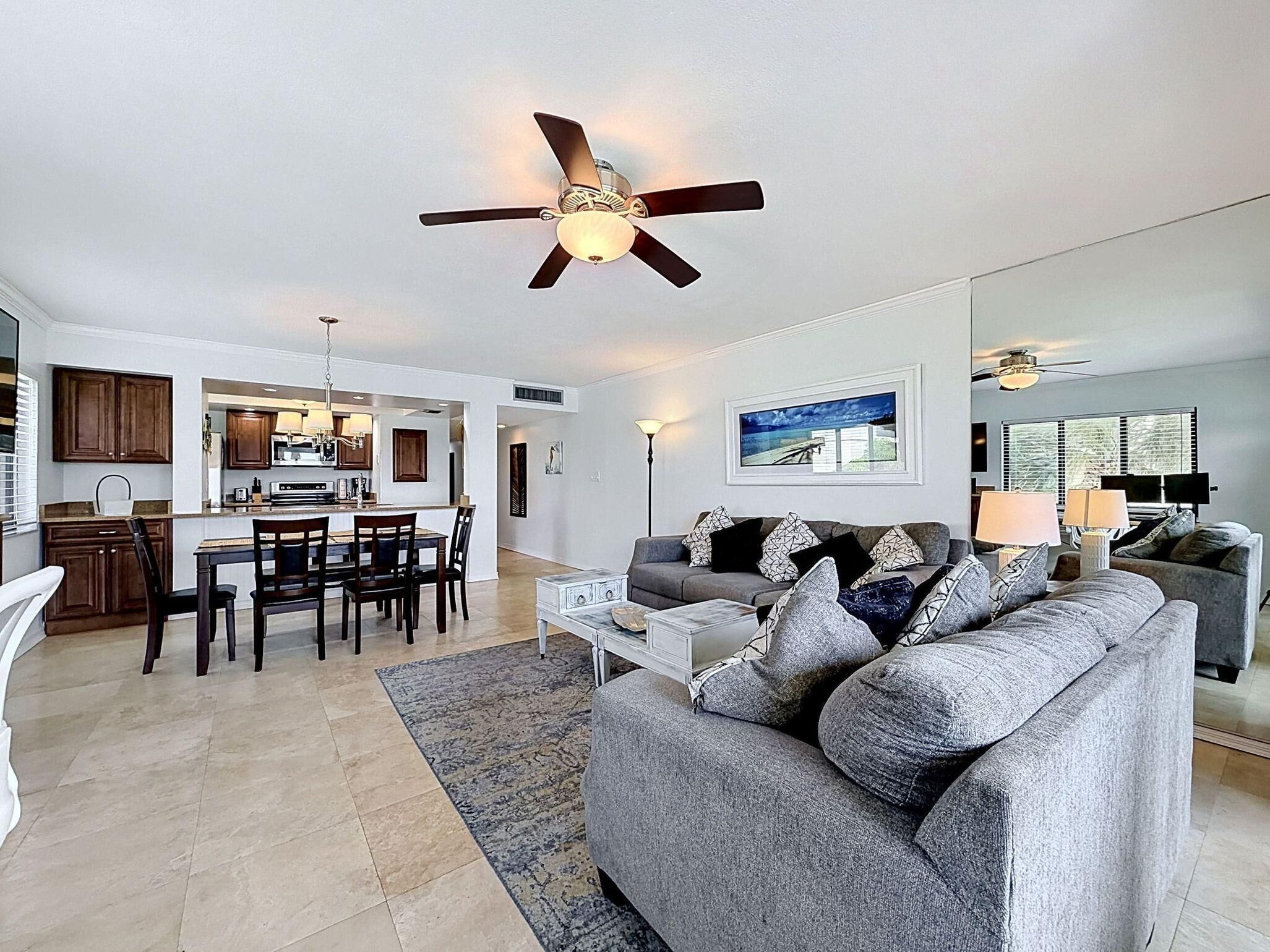 A modern living room with a grey sofa, a dining area with wooden chairs, a kitchen with dark cabinets, and a ceiling fan.