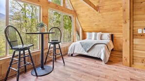 A cozy cabin room with a bed, two chairs, a table, and large windows offering a view of trees and mountains. - Amazing Views Hot Tub Game Room Pool (Gatlinburg)