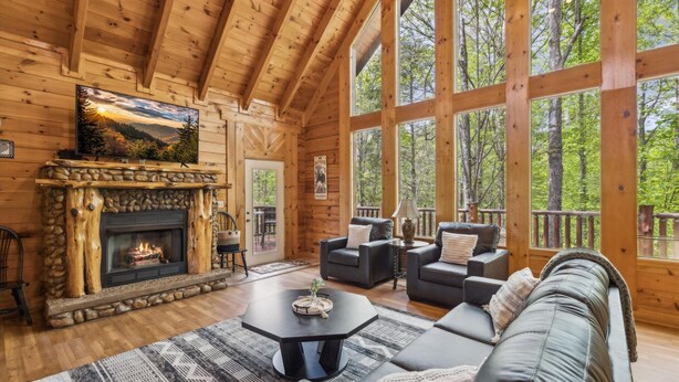 A cozy log cabin interior with a fireplace, comfortable seating, and large windows offering a view of the surrounding forest. - Amazing Views Hot Tub Game Room Pool (Gatlinburg)