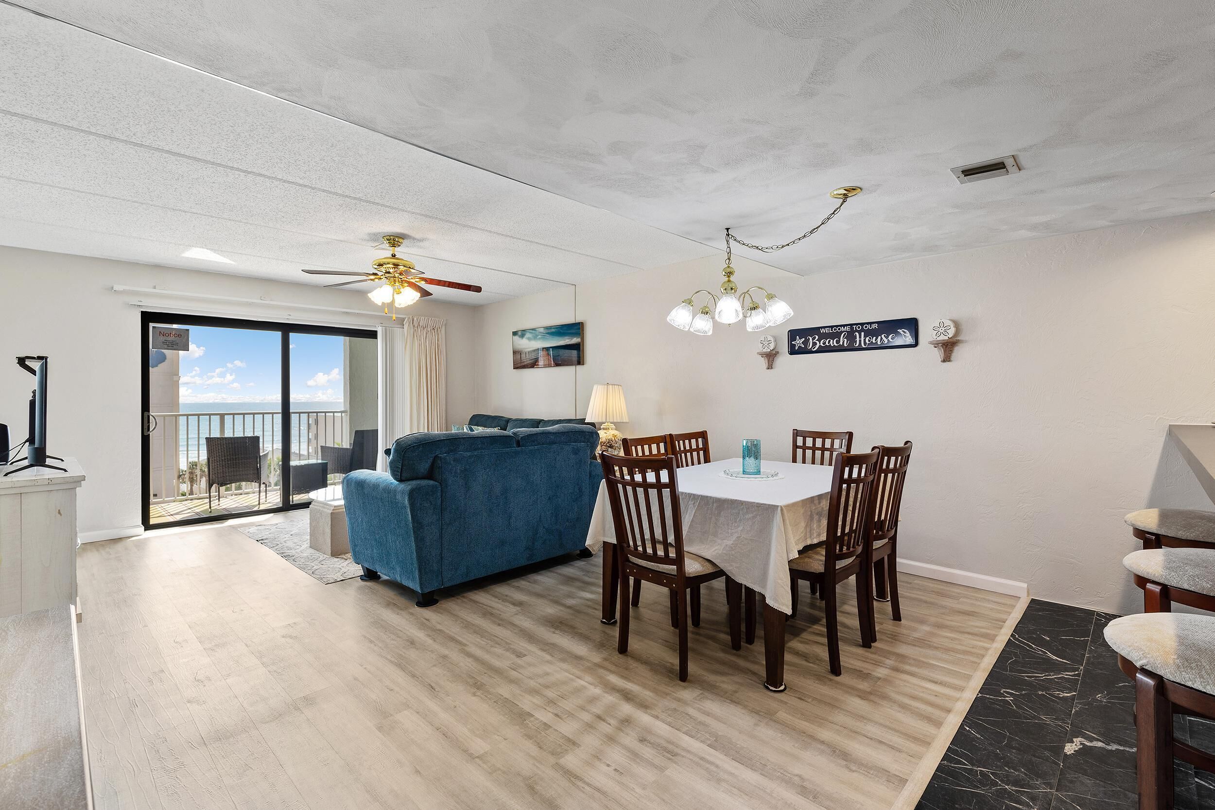 A modern living room with a blue sofa, dining table, and chairs, a ceiling fan, and a view through a sliding glass door to the outside.
