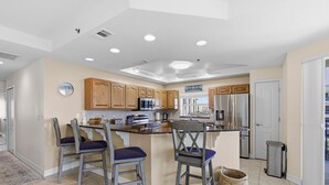 A kitchen with wooden cabinets, a stainless steel refrigerator, a microwave, and a sink. There are two bar stools with blue cushions.