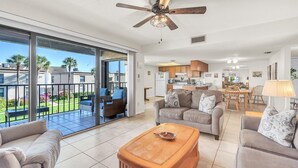 A spacious living area with a balcony, a ceiling fan, and a kitchen in the background.