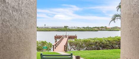 Vista de un lago con un muelle de madera que lleva a un embarcadero, un banco verde y un letrero que indica una zona de parque.