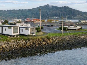 A waterfront RV park with several RVs and trailers, a rocky shoreline, and a backdrop of hills and buildings.
