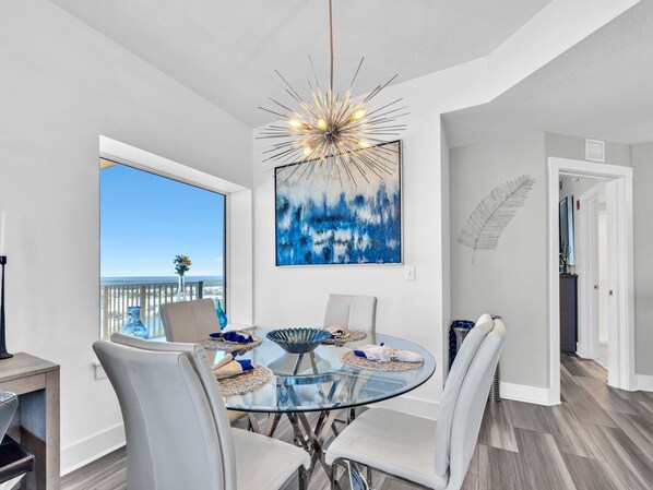 A dining area with a glass table, white chairs, a modern chandelier, and a large window with a view of the beach.