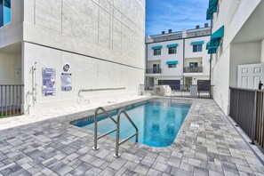 Una piscina con una escalera de metal, rodeada por un suelo de baldosas y un edificio con balcones.