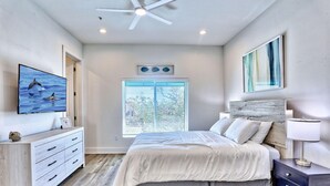 Une chambre à coucher comprenant un lit, une commode, une table de chevet, un ventilateur de plafond, une fenêtre et un tableau accroché au mur.