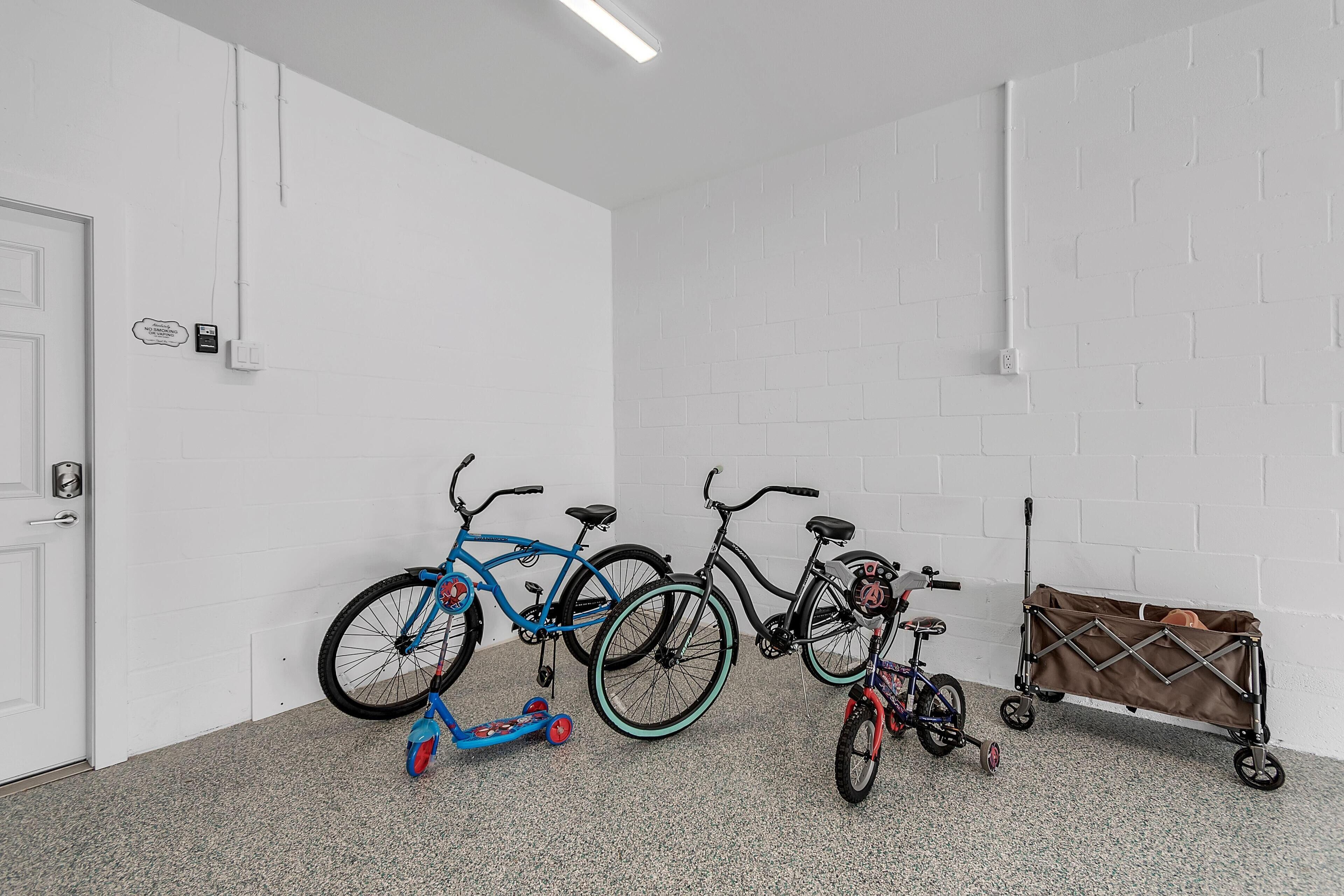 Une pièce aux murs en briques blanches, avec une porte blanche et un sol gris à motifs. Trois vélos et une poussette sont appuyés contre le mur.