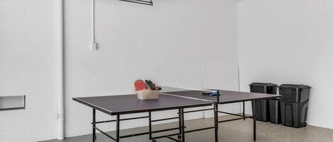 A ping pong table in a room with white brick walls, a white ceiling, and a grey floor. There are two trash bins against the wall.