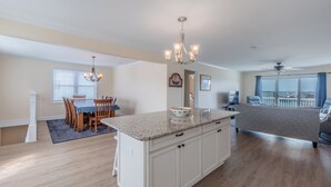 A spacious living area with a large island, a dining area with wooden chairs, and a balcony with a view. - Bear's Den by Topsail Realty (Holly Ridge)