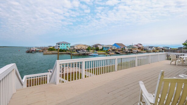 A waterfront property with a wooden deck, white railings, and a view of colorful houses across the water. - Bear's Den by Topsail Realty (Holly Ridge)
