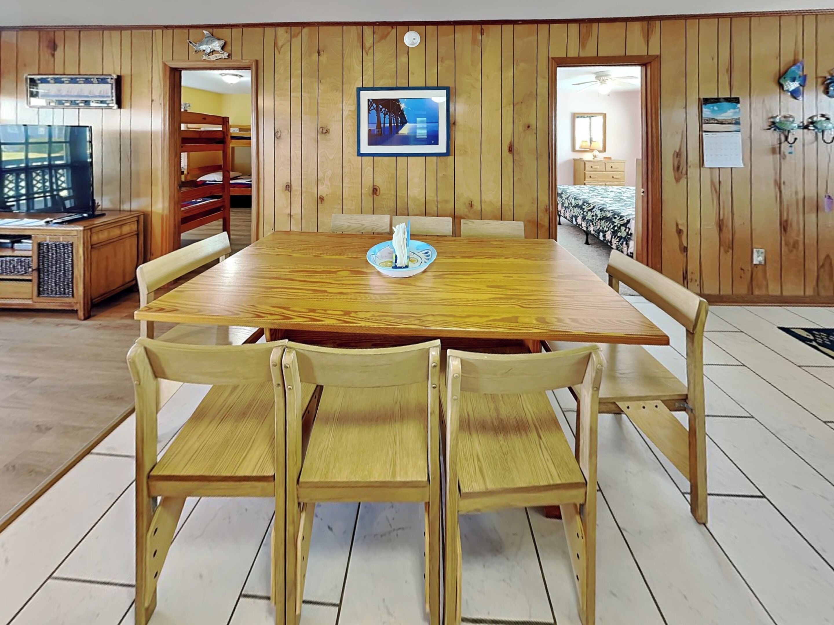 A wooden-paneled room with a round table and six chairs, a television, and a framed picture on the wall.