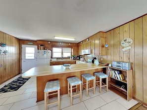 A kitchen with wooden cabinets, a refrigerator, a microwave, and a coffee maker. There are three bar stools and a bookshelf with books. - Boyd by Topsail Realty (Holly Ridge)