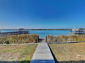 A wooden pier leading to a dock with a small shelter, surrounded by grassy areas and a calm body of water.