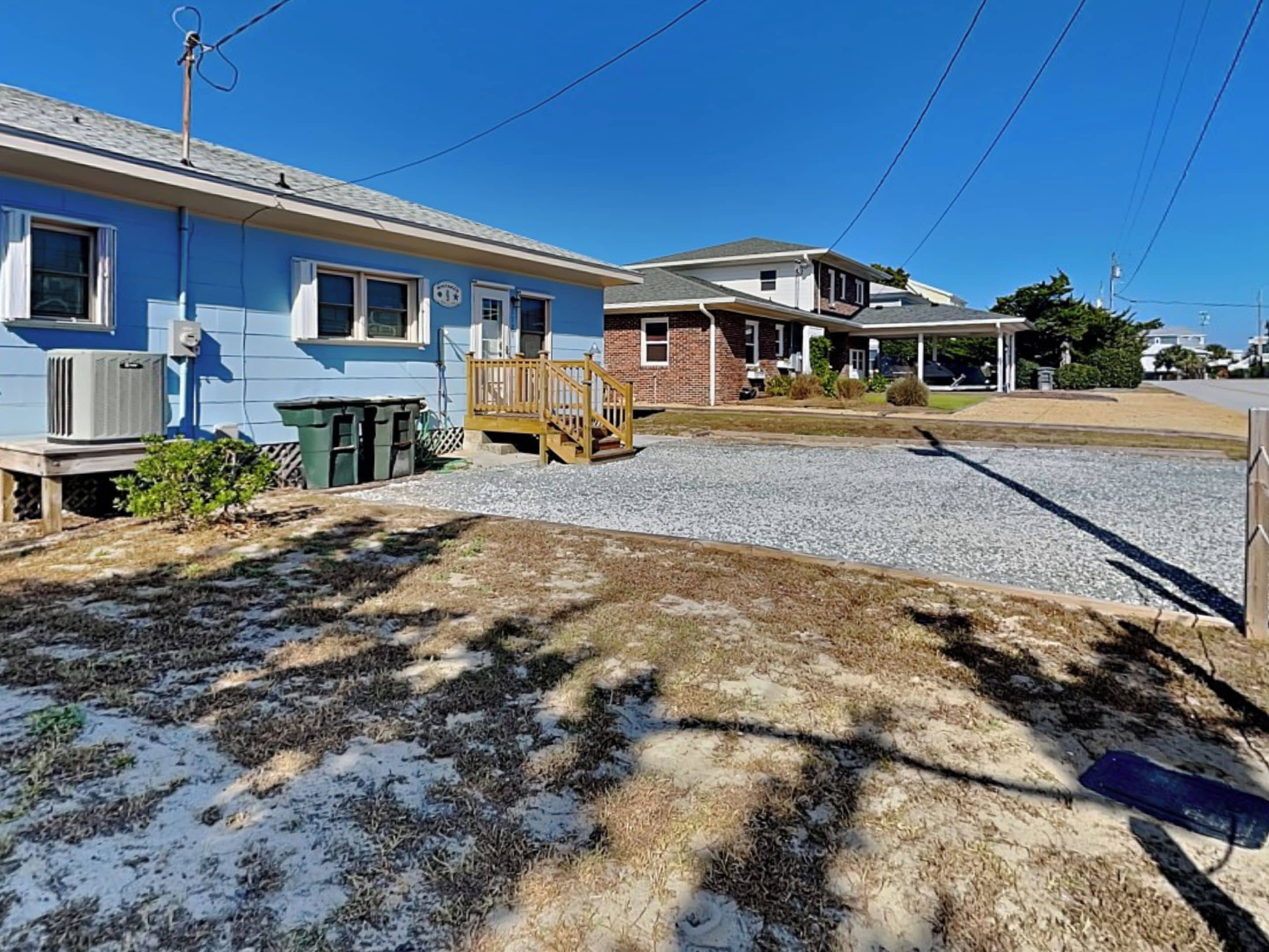 A residential area with two houses, one with a blue exterior and the other with a brick facade. There is a gravel driveway in front of the houses, and a wooden deck attached to the blue house.