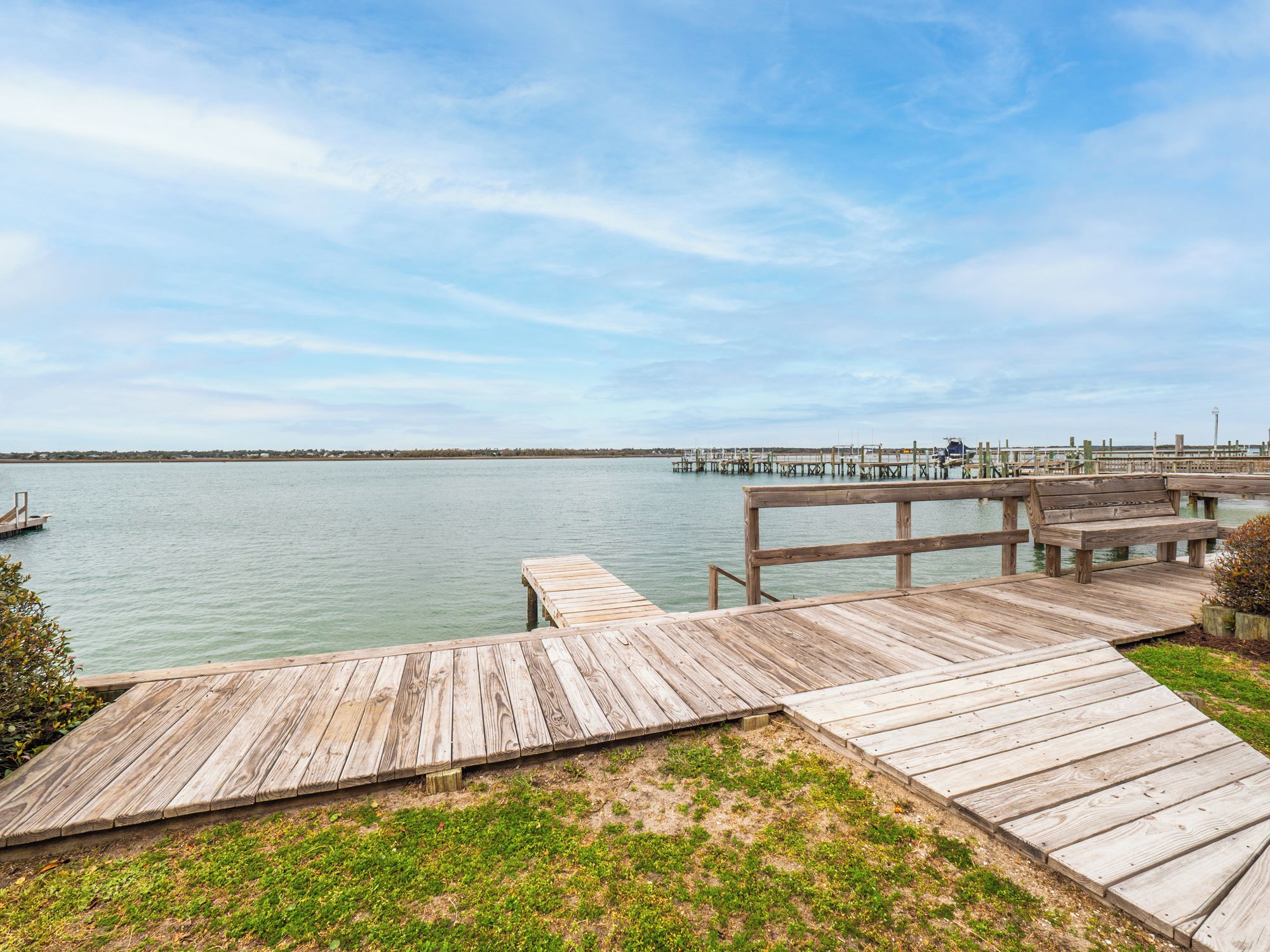 A wooden pier extending into calm water, with a grassy area in the foreground and a distant shoreline.