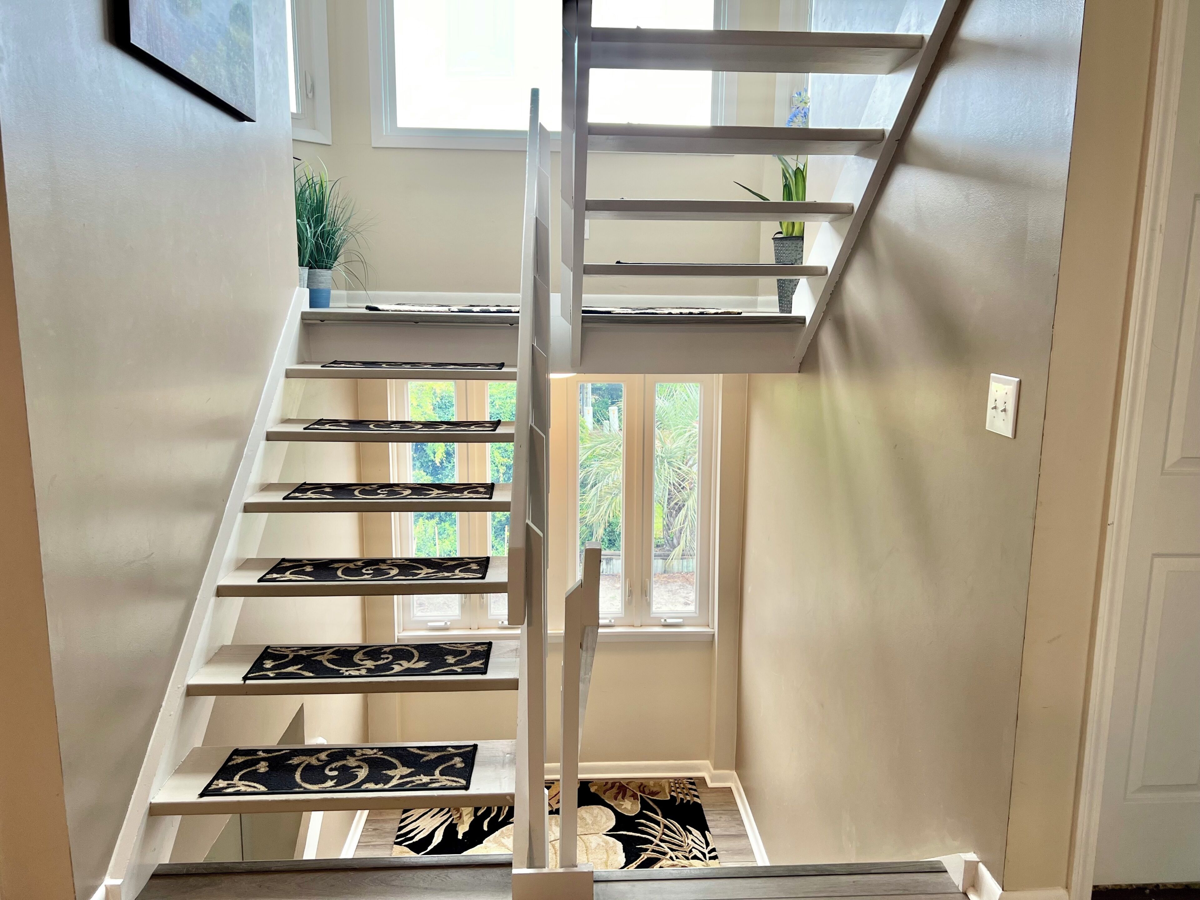 A staircase with white railings and black patterned mats, leading to a landing with a window view of greenery.
