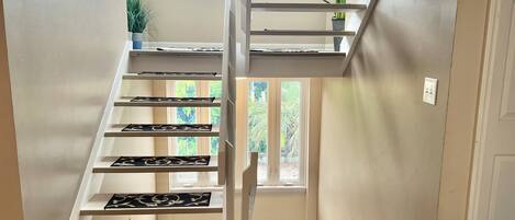 A staircase with white railings and black patterned mats, leading to a landing with a window view of greenery.