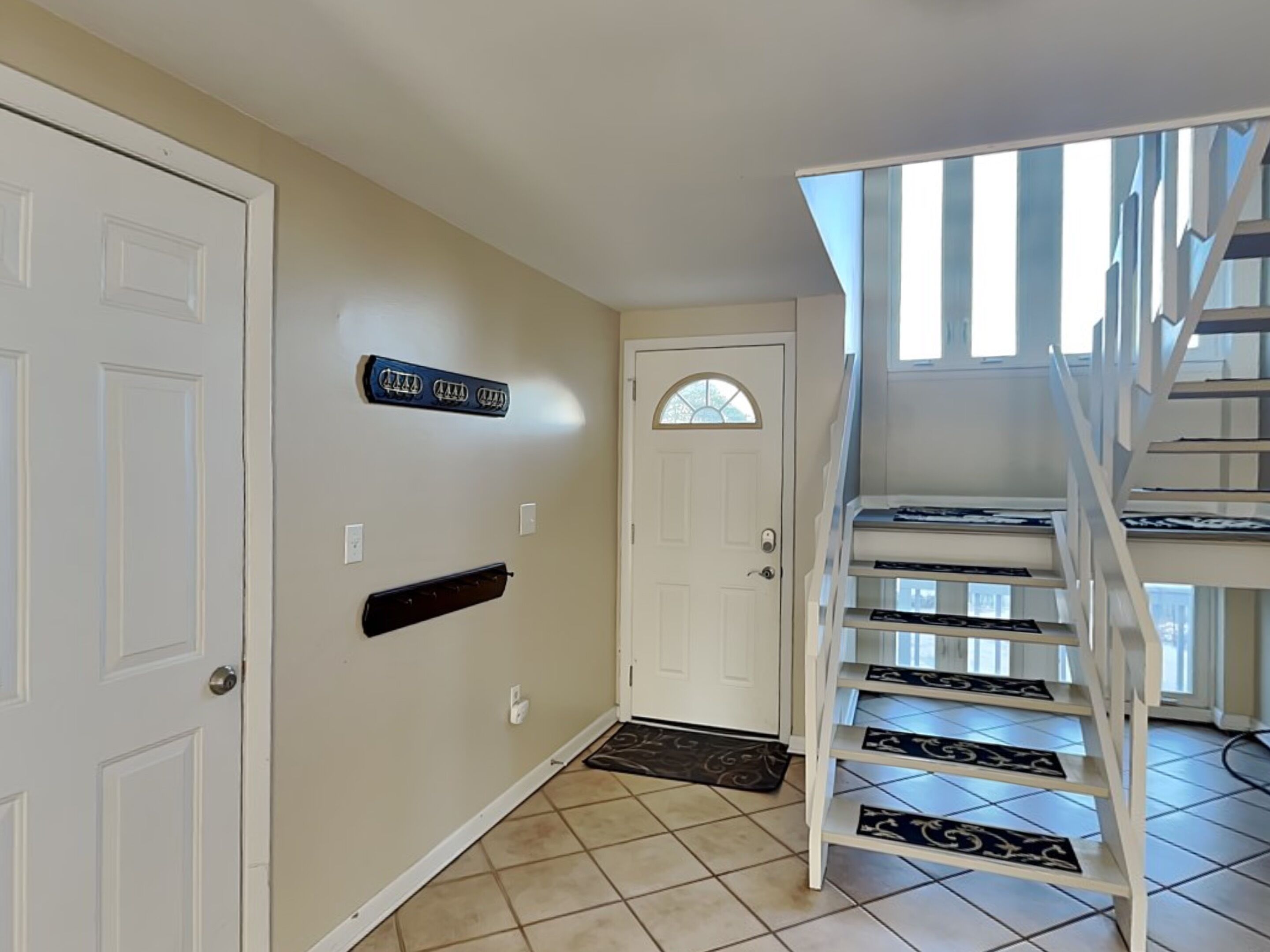 Un couloir aux murs de couleur beige, une porte blanche, un porte-manteau noir fixé au mur et un escalier blanc avec des marches noires.
