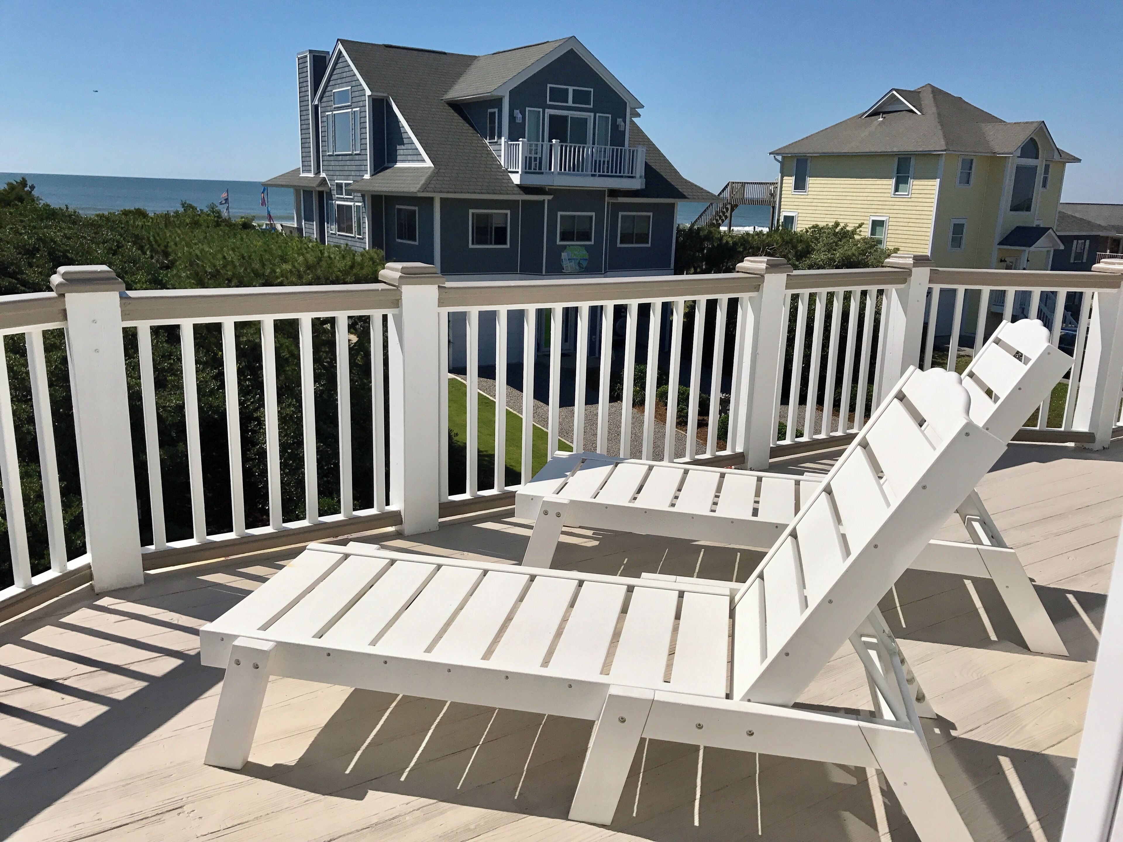 A wooden deck with white railings, a white lounge chair, and a view of a large house with a balcony.