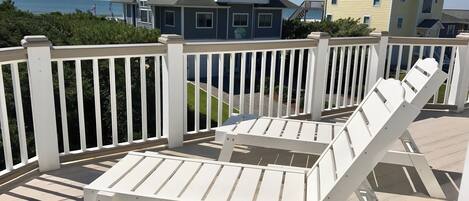 A wooden deck with white railings, a white lounge chair, and a view of a large house with a balcony.