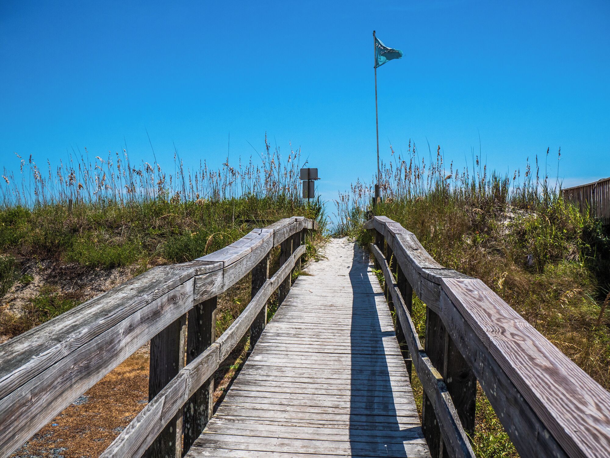 A wooden boardwalk leading to a flagpole with a flag, surrounded by tall grasses and a clear blue sky.