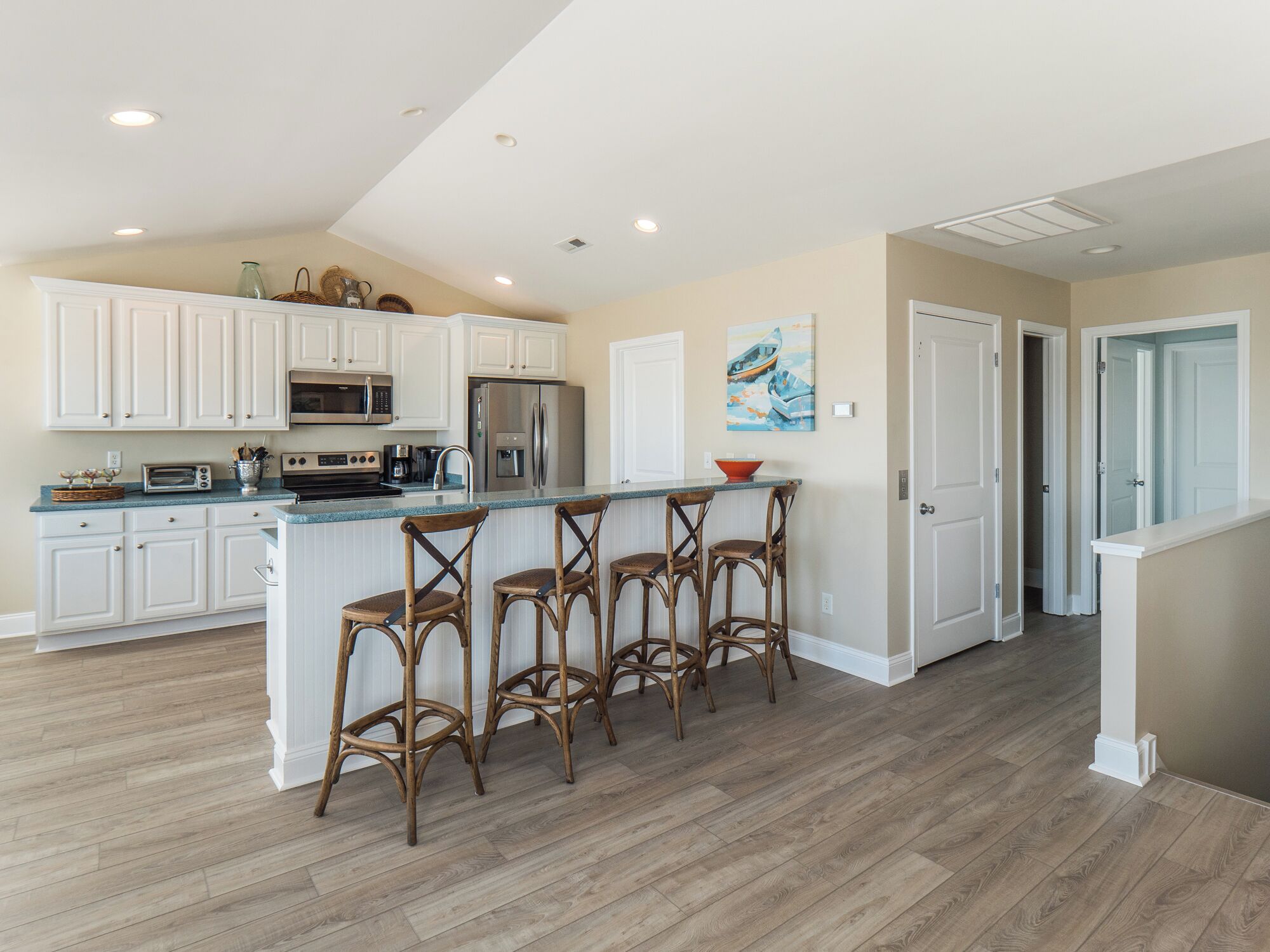 A modern kitchen with white cabinets, a stainless steel refrigerator, a microwave, and a toaster. There are four wooden bar stools at the island.
