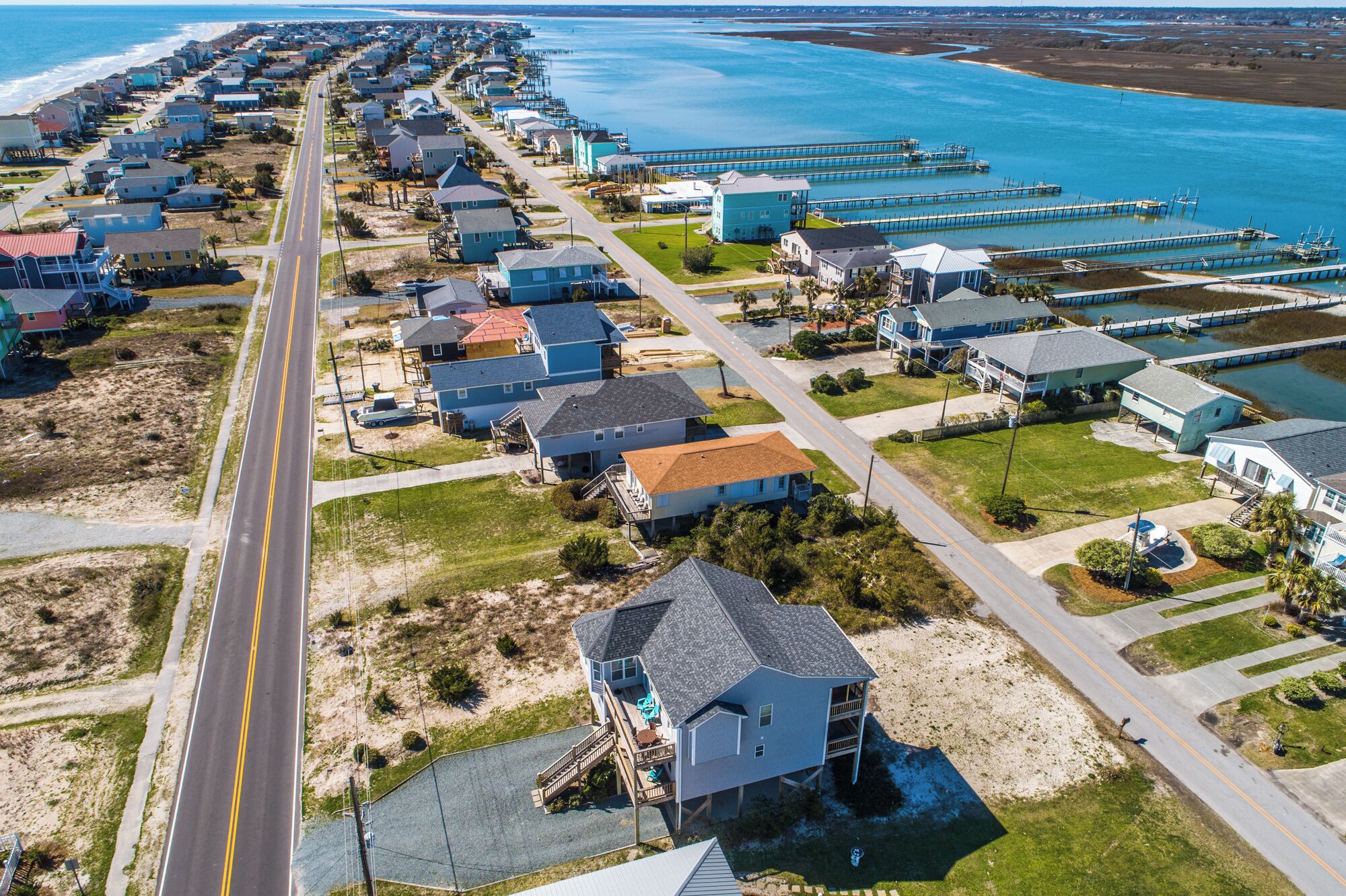 An aerial view of a coastal residential area with houses, a road, and a pier extending into the water.