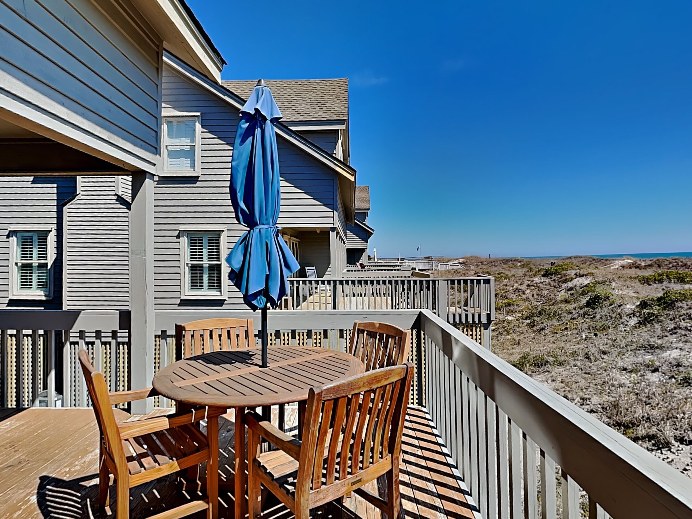 Une terrasse en bois avec une table et des chaises, un parasol bleu et une vue sur la plage et l’océan.