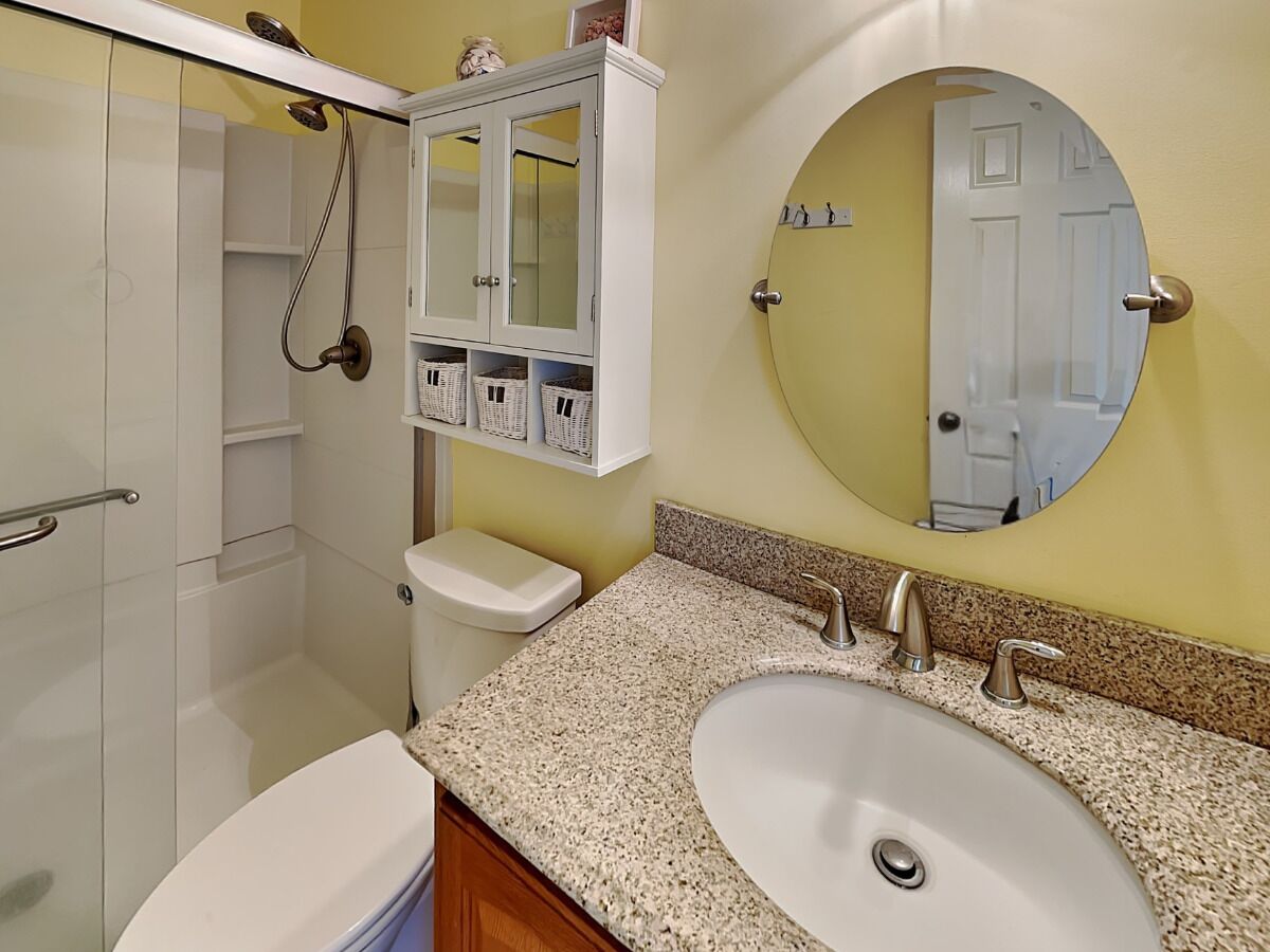 A bathroom with a white toilet, a granite countertop sink, a mirror, and a cabinet with baskets.